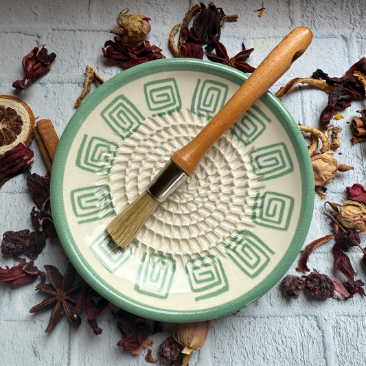 Ceramic bowl with decorative patterns, a wooden brush, and dried herbs on a textured surface.
