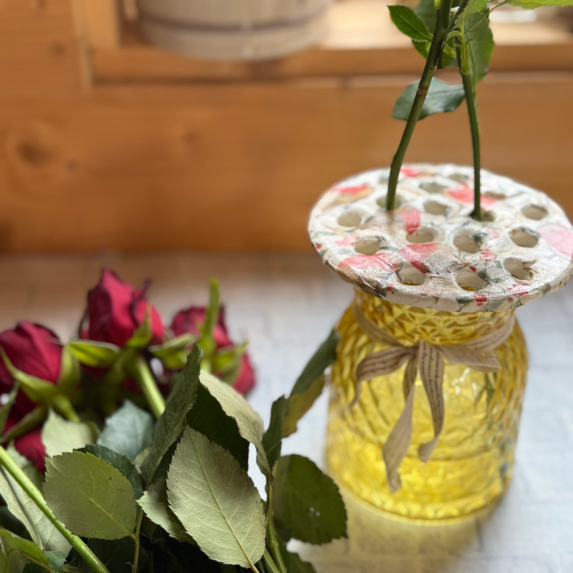 Yellow glass bottle with floral flower arranging frog holding a rose, surrounded by red roses on a wooden surface.