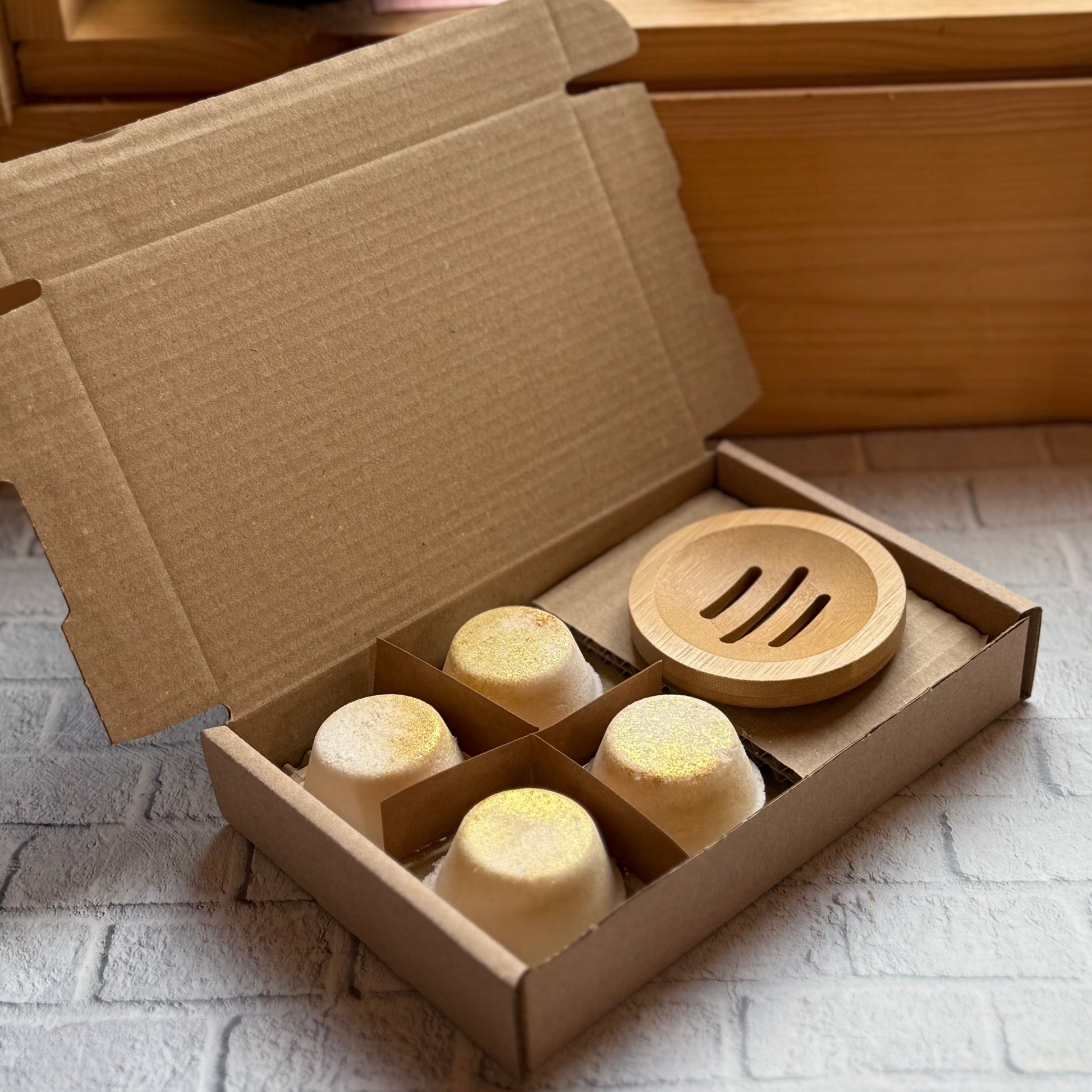 Gift box of 4 aromatherapy shower steamers and bamboo dish on a stone surface with a wooden cabinet in the background.